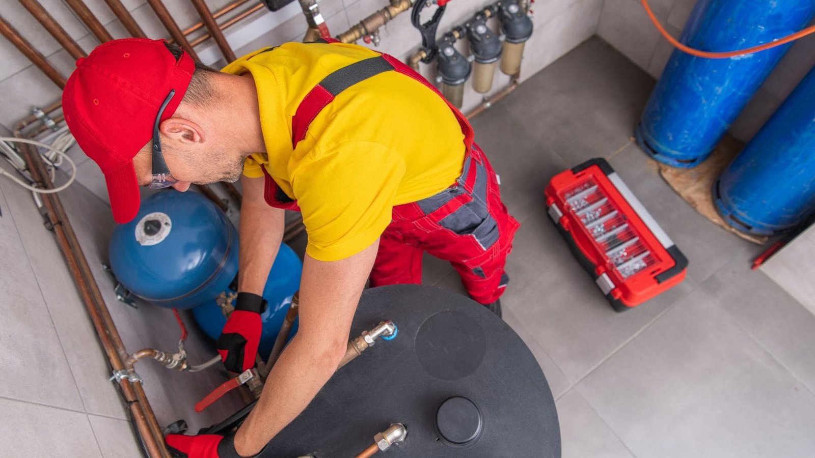 Indoor vs outdoor tankless water heater maintenance worker in red overalls inspecting plumbing connections.