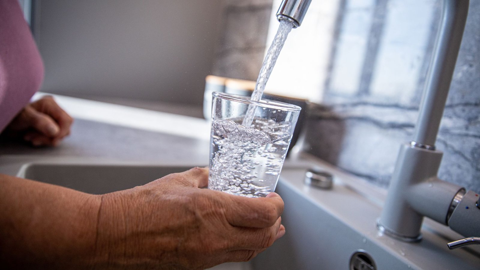 Chloramine removal process illustrated by an older hand filling a glass from a modern kitchen faucet.