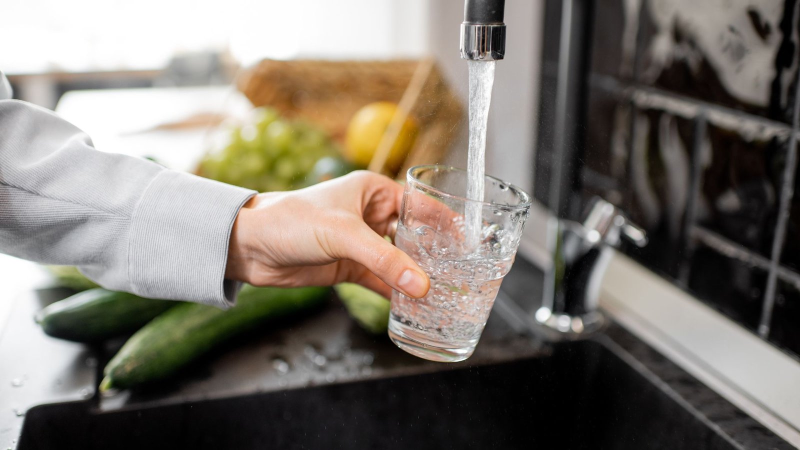 Tannins clean tap water flowing into glass in modern kitchen with fresh produce on the counter.