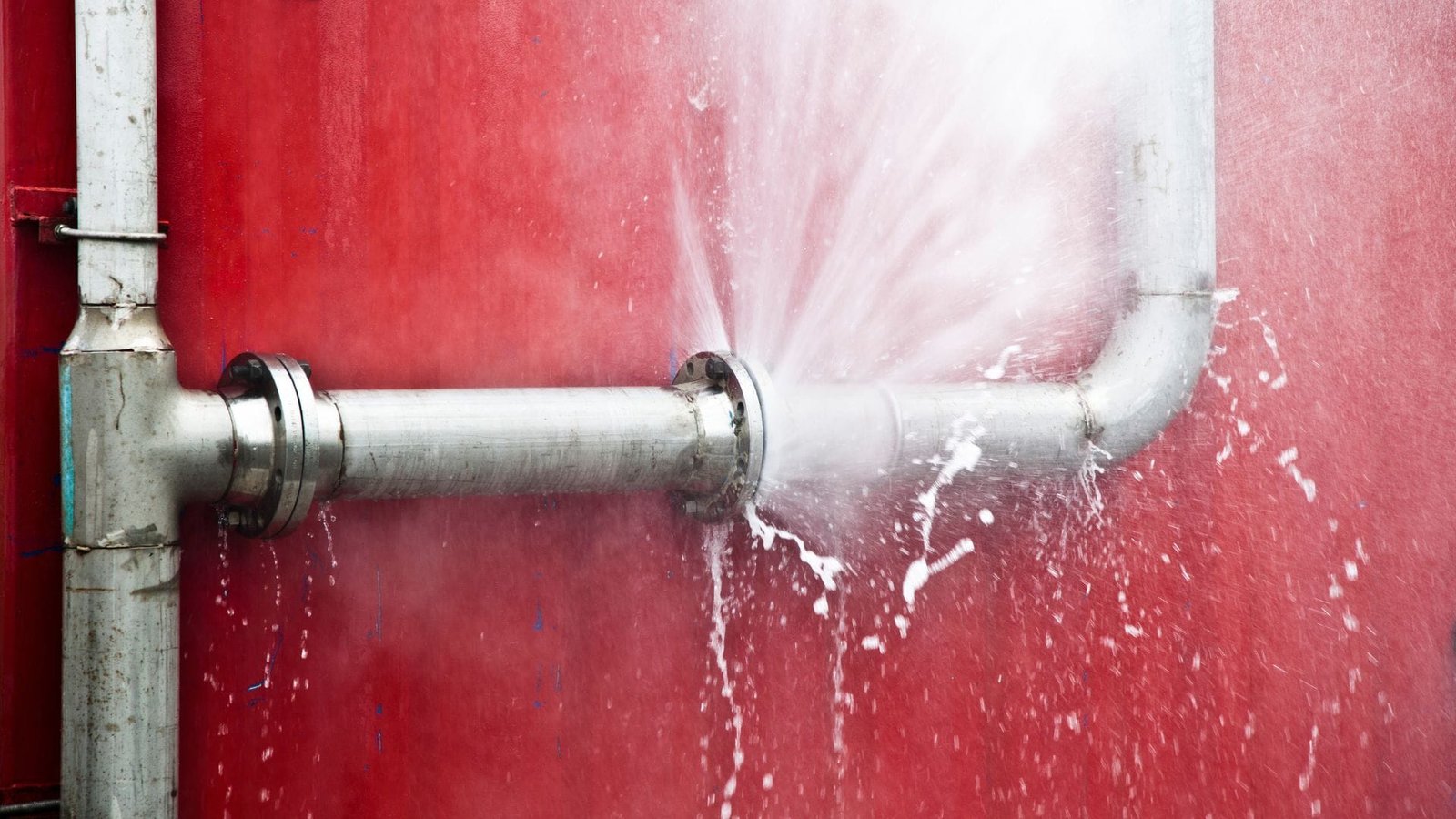Water softener leaking burst pipe with high-pressure water spraying against red industrial background.
